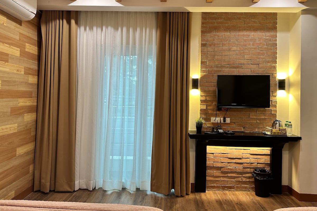 A modern room with wooden flooring, a large window with sheer curtains, a brick accent wall, a mounted TV, and a small table with a trash bin.