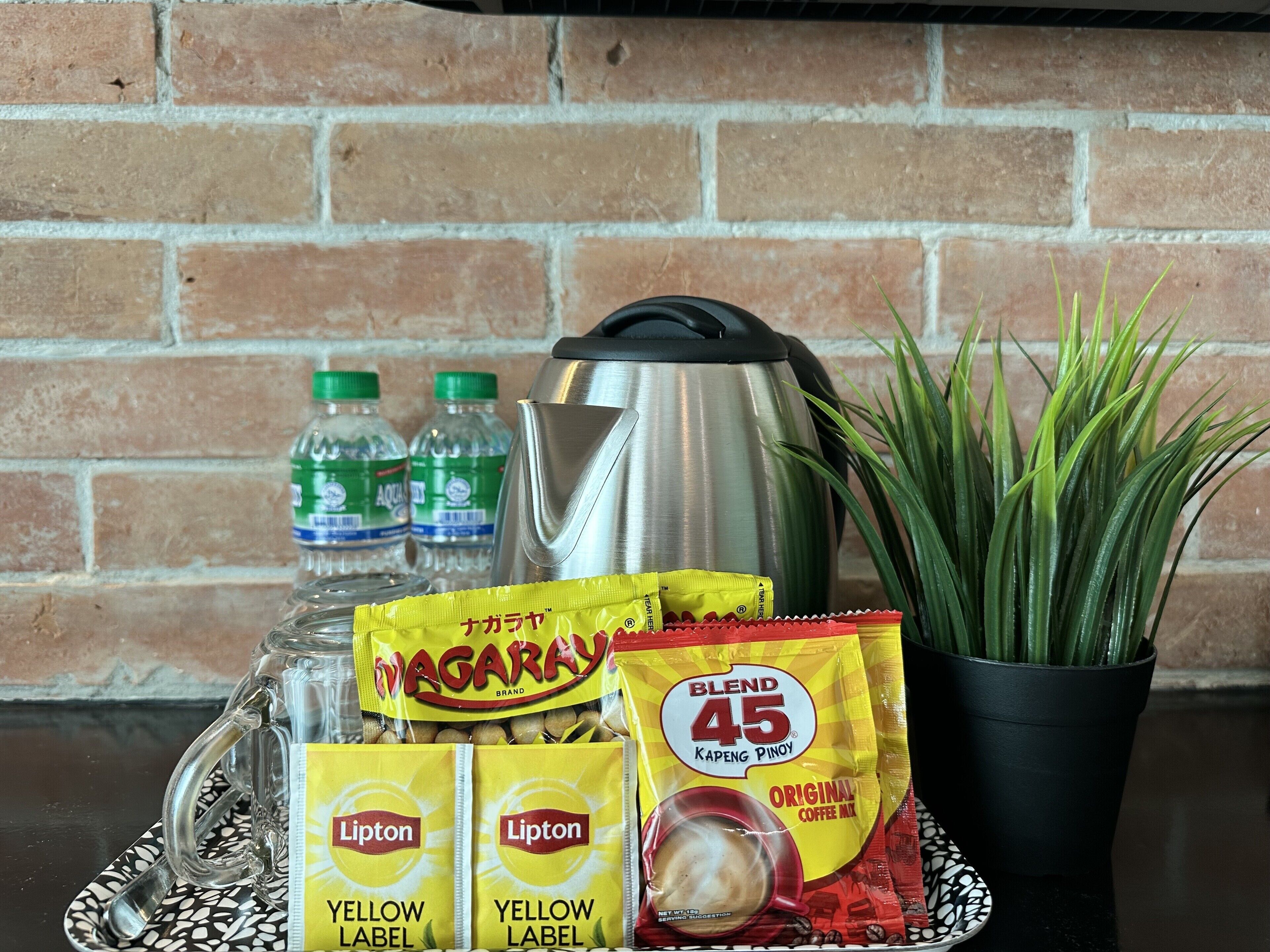 A coffee-making setup with Lipton Yellow Label coffee packets, a Blend 45 Kapeng Pinoy coffee mix, a stainless steel kettle, and two water bottles.