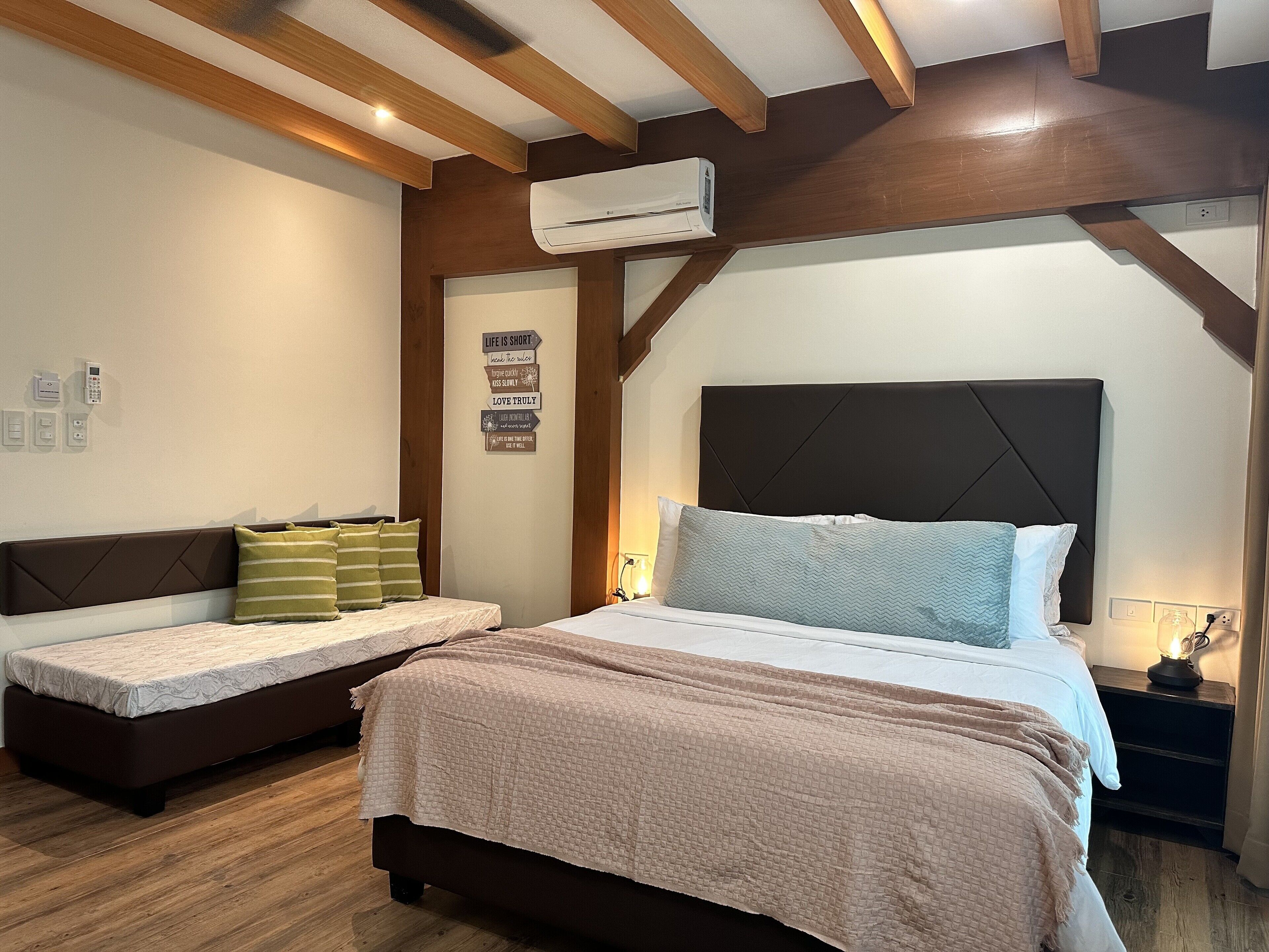 Une chambre avec un grand lit, un petit canapé, des poutres en bois au plafond, un climatiseur et une citation inspirante sur le mur.