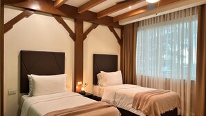 Habitación de hotel con dos camas, vigas de madera en el techo, ventilador de techo y una ventana con cortinas translúcidas.
