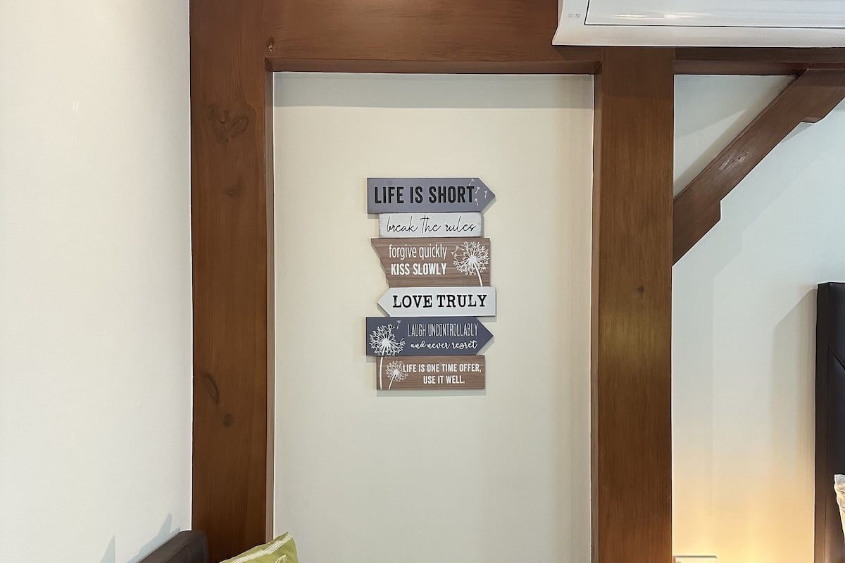 A bedroom with a bed, pillows, a wall-mounted sign with life advice, an air conditioner, and a bedside lamp.