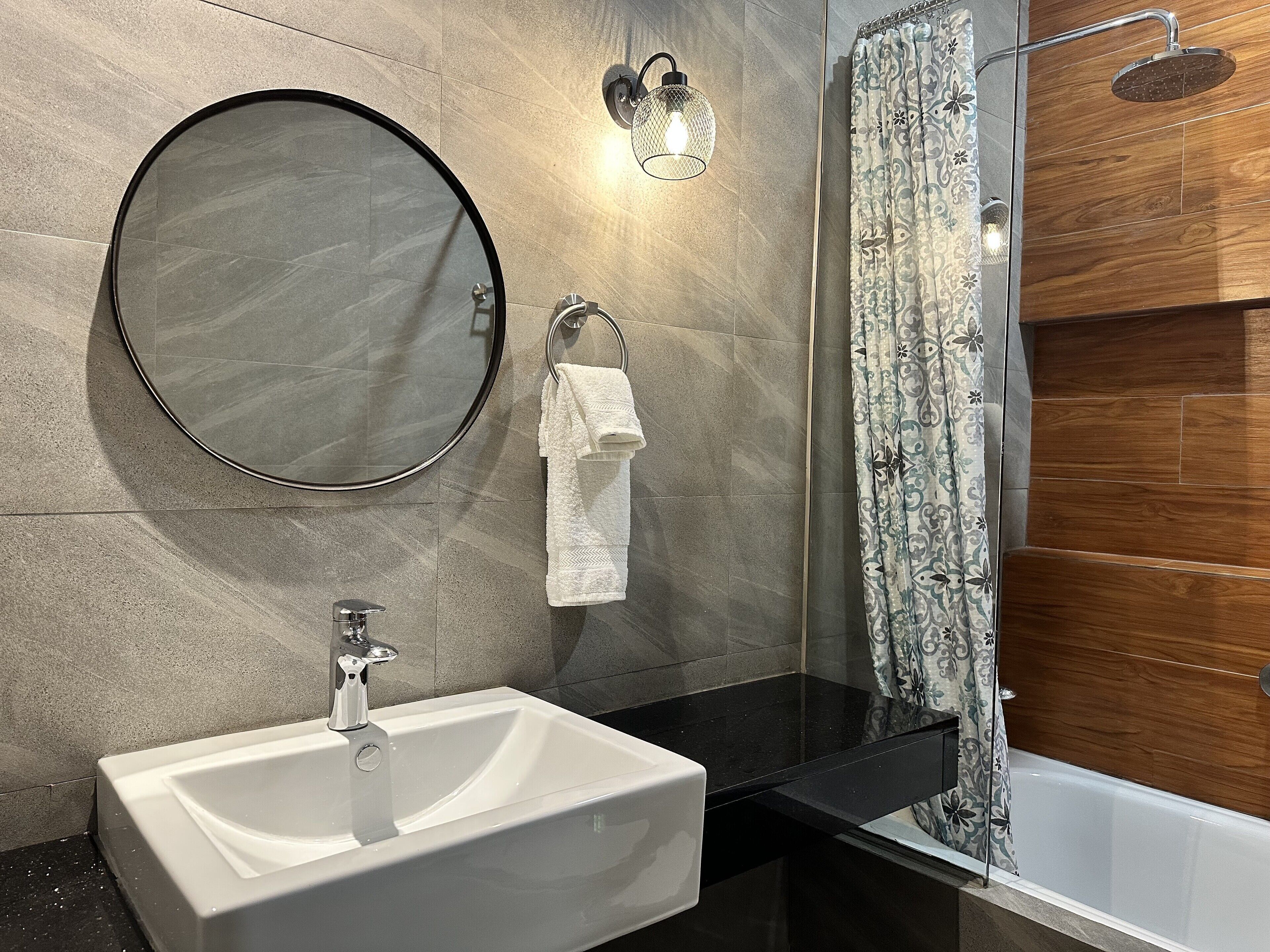 Un bagno moderno con uno specchio rotondo, un lavandino bianco e una doccia con parete rivestita in legno.