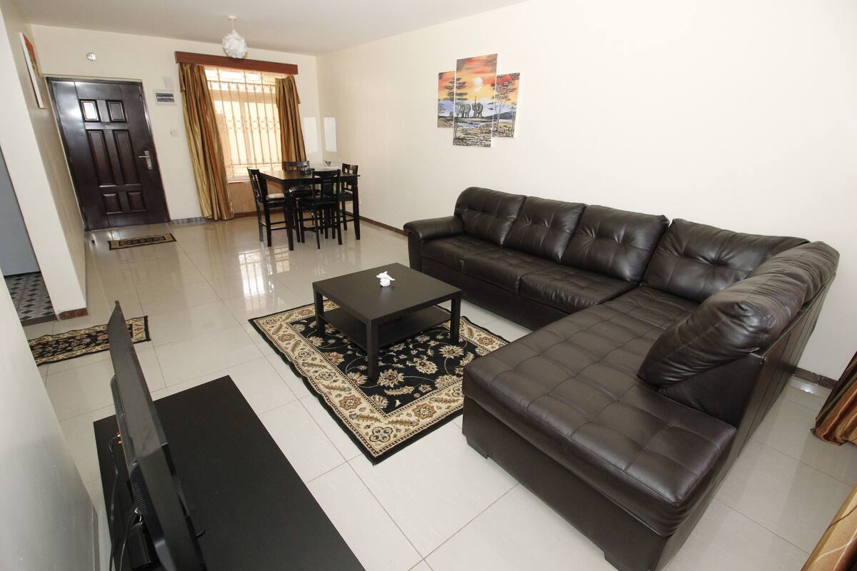 A living room with a brown leather sofa, a black coffee table, a TV, and a dining area with chairs and a table.