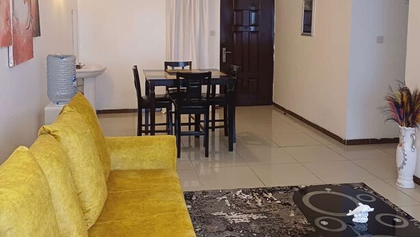 Una sala de estar con un sofá amarillo, una mesa de comedor con sillas, un dispensador de agua y una puerta que da a otra habitación.