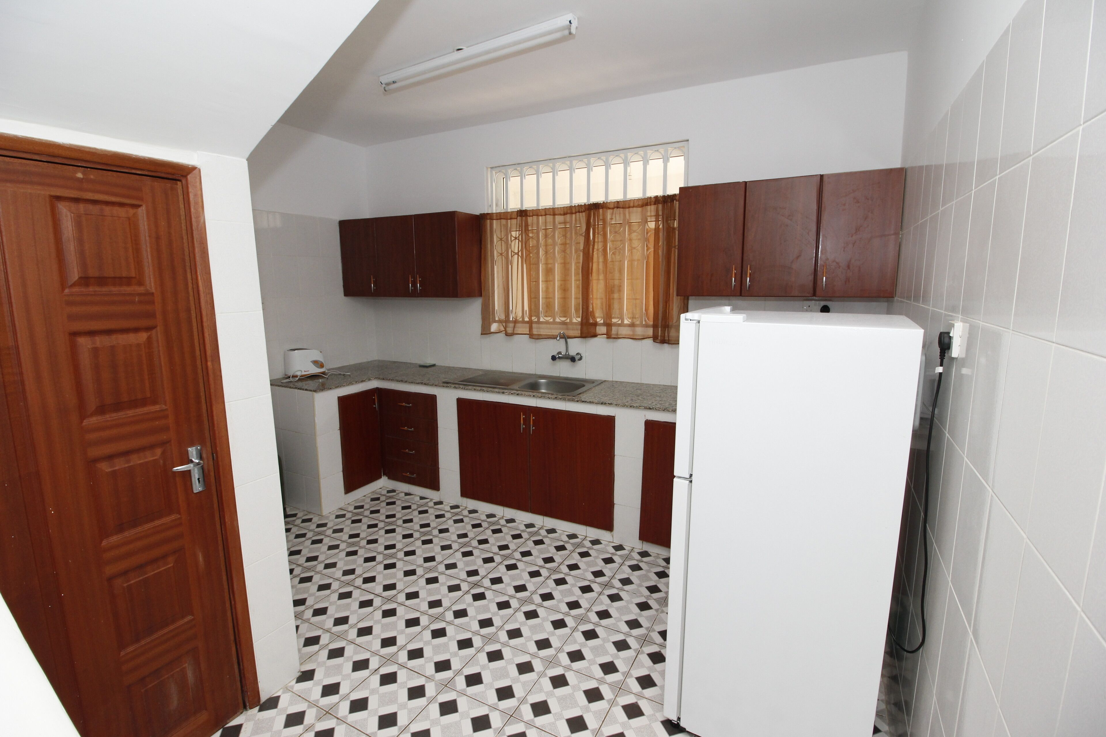 Una cocina compacta con refrigerador, gabinetes de madera, una ventana con cortinas y piso de baldosas.