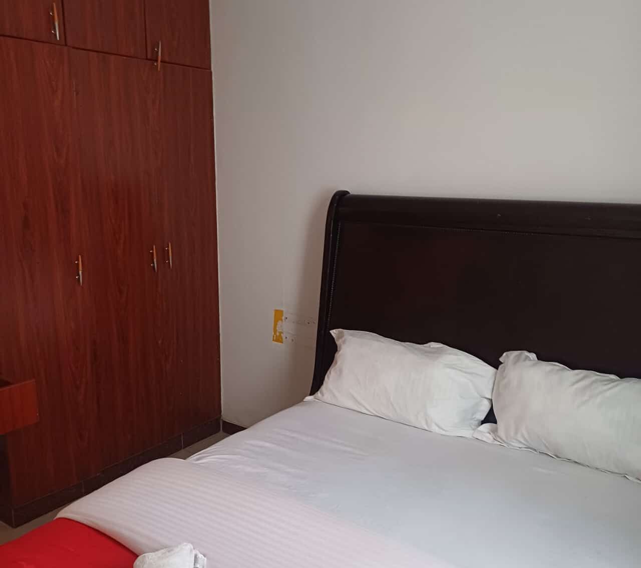 A bedroom with a bed, white pillows, and a red blanket. There is a wooden wardrobe with two doors and a small red table.