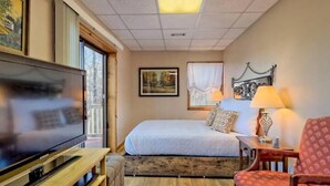 Une chambre d’hôtel avec un lit, un téléviseur posé sur un meuble en bois, une fenêtre avec des rideaux et une lampe sur une table de chevet.