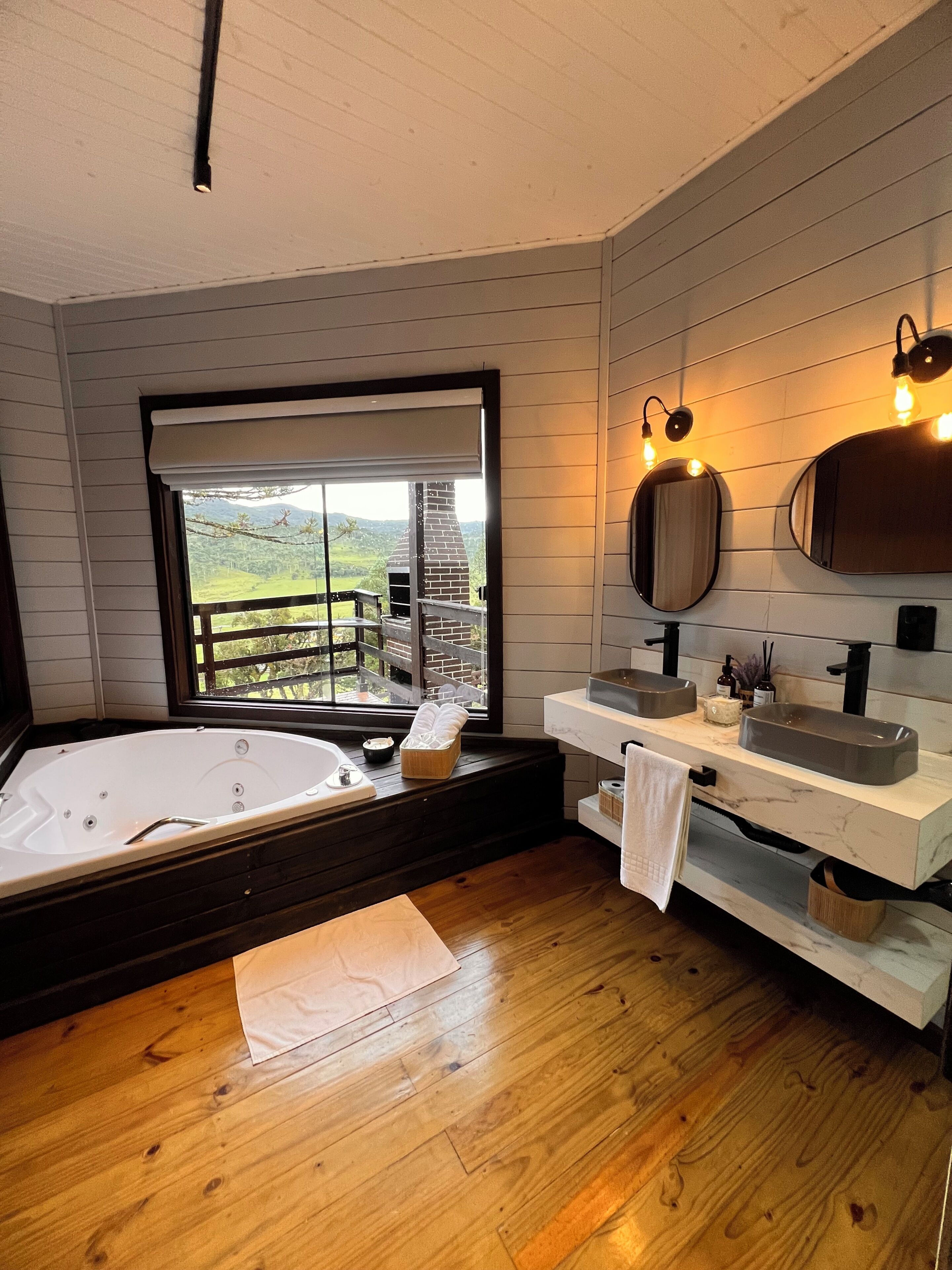 A modern bathroom with a large bathtub, dual sinks, and a view of a green landscape through a window.