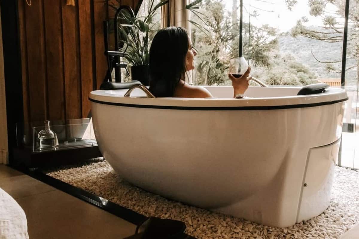Une personne se détend dans une baignoire sur pied, un verre de vin à la main, avec une plante en pot à proximité et une vue sur des arbres et des collines.