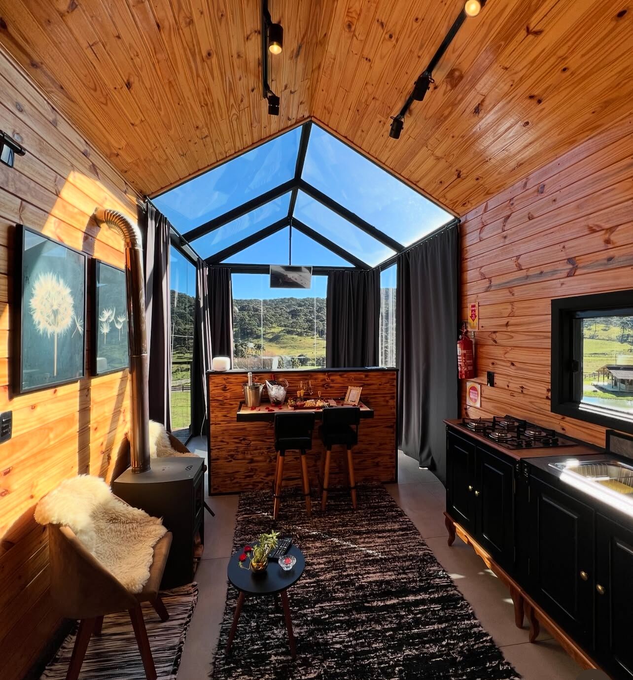 A cozy cabin interior with a wooden interior, a dining area, a fireplace, and a large window offering a scenic view.