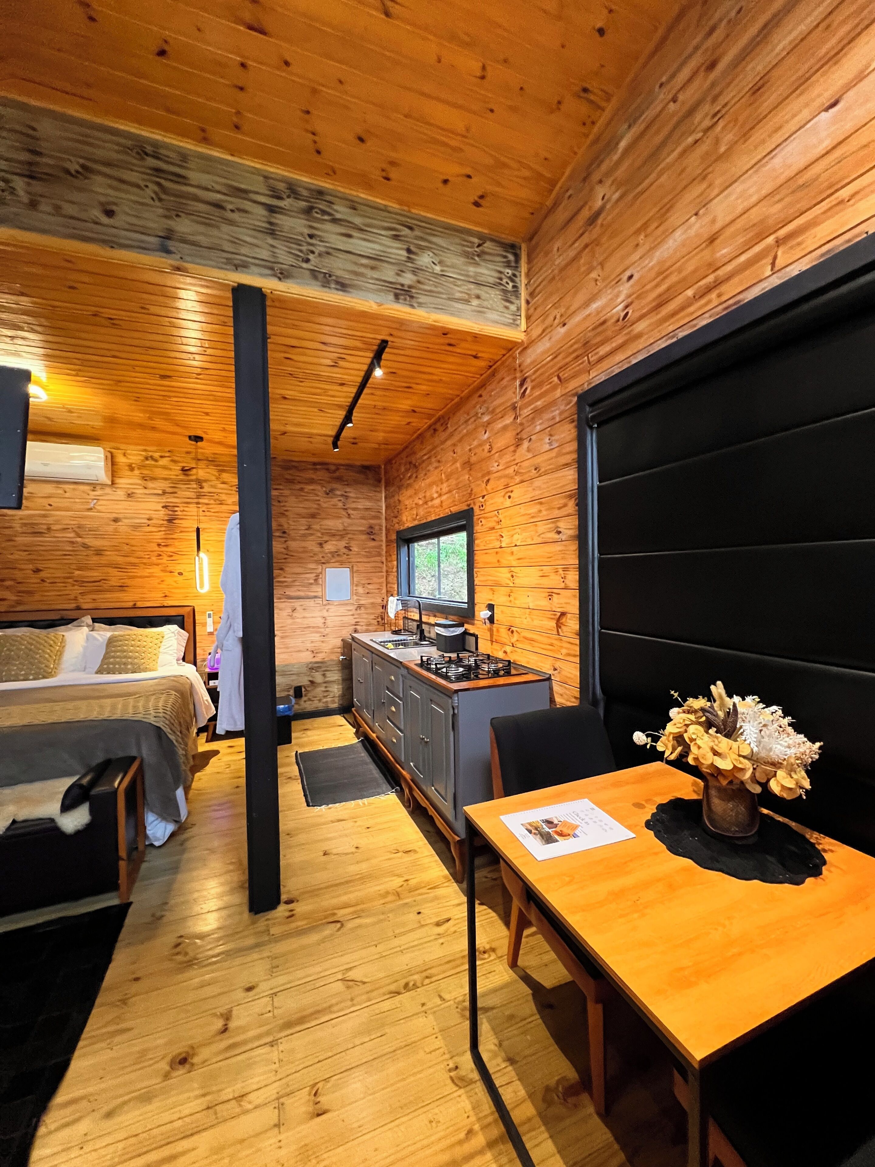 A wooden interior with a bed, kitchenette, dining table, and a large window.