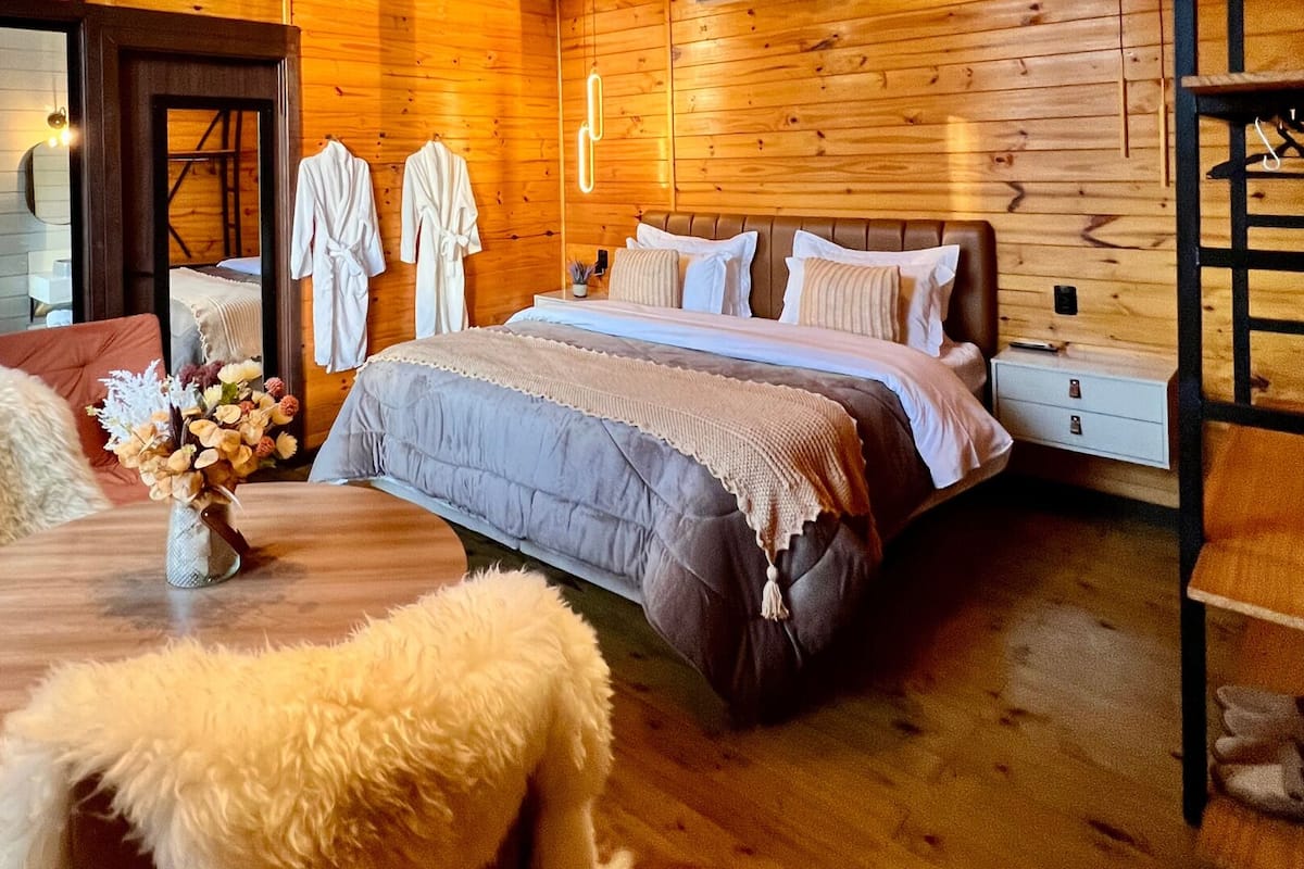 Une chambre confortable dans un chalet en bois, avec un lit, une table ornée d’un vase de fleurs et un adorable chien blanc à la fourrure douce.