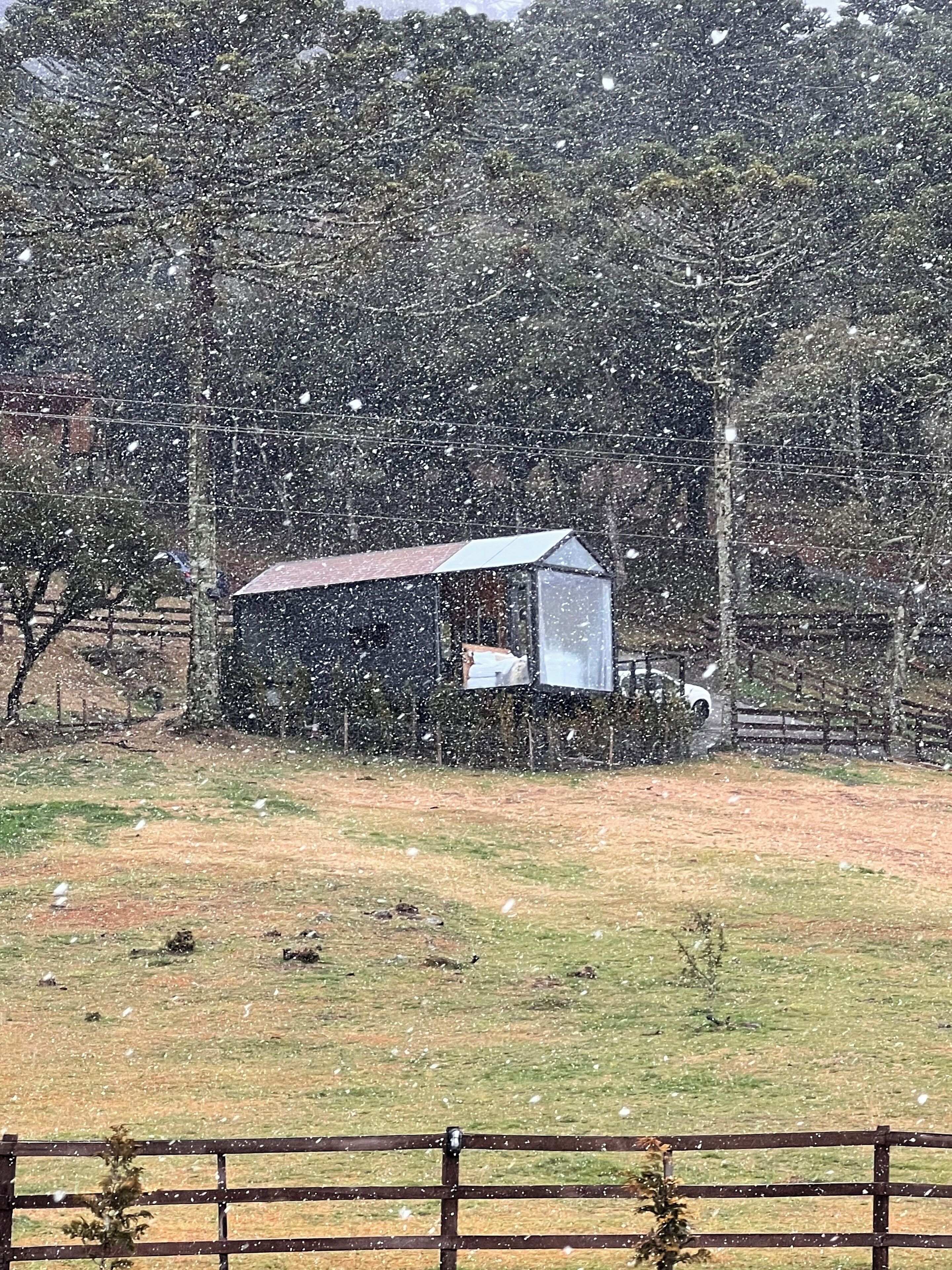 A small, single-story building with a sloped roof, surrounded by a fenced area and trees.