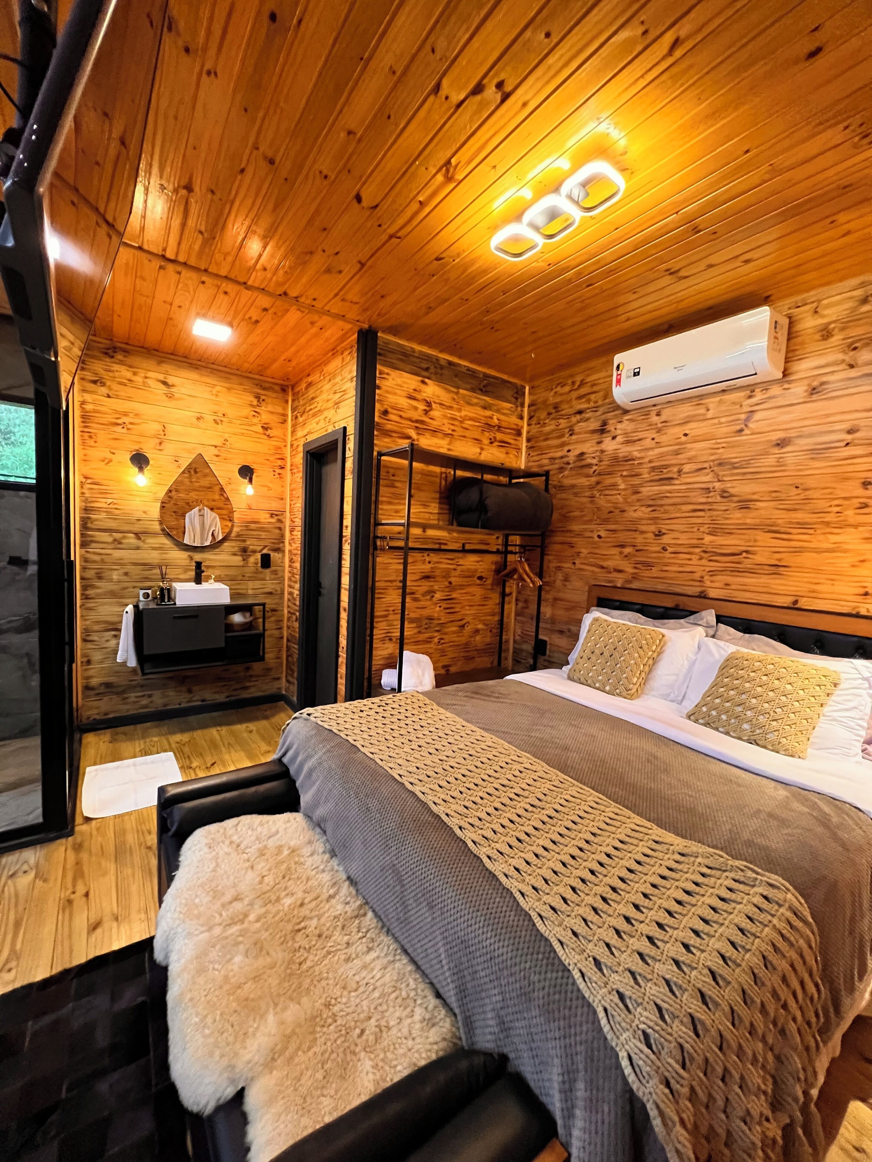 Interior de una cabaña de madera con cama, espejo, lavabo y lámpara en el techo.