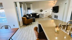 Una cocina moderna con una isla central, taburetes de bar, una mesa de comedor y una sala de estar con sofá y televisor.