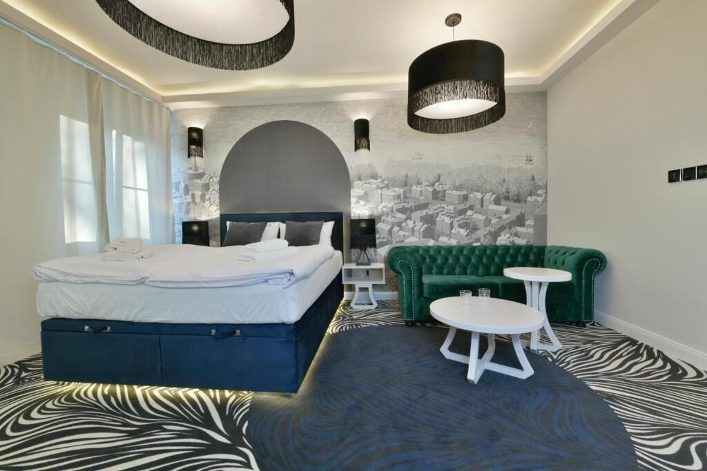 Una habitación de hotel moderna con una cama grande, un sofá verde, dos mesitas pequeñas y un mural con un paisaje urbano en la pared.