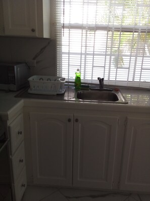 A kitchen with white cabinets, a microwave, a dish rack, and a window with blinds. - Garden Apartment modern clean and well kept, comfortable cool burglar bars nets (Mount Standfast)