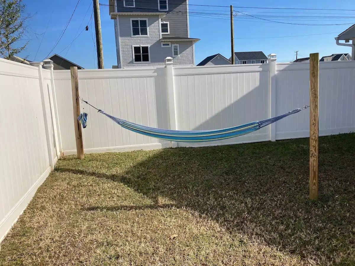 Un hamac est suspendu entre deux poteaux en bois dans une cour arrière clôturée, où l’herbe est sèche.