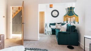 Een woonkamer met een groene bank, een klok aan de muur en een trap die naar een andere kamer leidt.