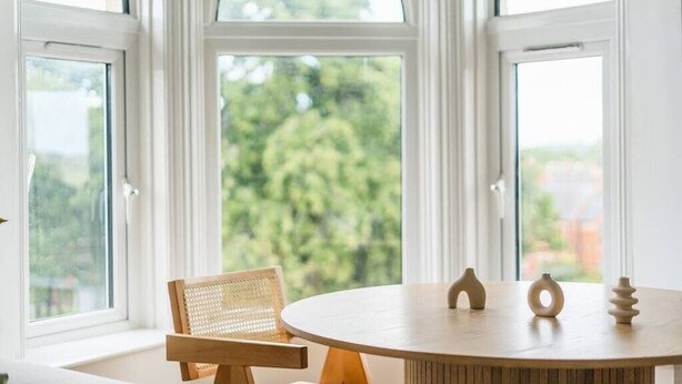 Een ronde houten tafel met drie decoratieve objecten, een rieten stoel en zicht op groen door grote ramen.