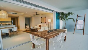 Una zona de menjador moderna amb una taula de fusta i cadires blanques, una cuina al fons i una planta gran en test.