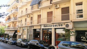 Um edifício de vários andares com varandas, uma loja chamada "Hadjopoulos" e carros estacionados na rua.