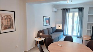 Una sala de estar moderna con un sofá gris, una mesa redonda de madera y una mesita auxiliar con una lámpara. Hay un cuadro en la pared y una ventana con cortinas.