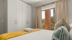 A bedroom with a bed, a wooden headboard, a window with curtains, and a brick wall visible through the window. - Comfort Living In The Heart Of Century City (Cape Town)