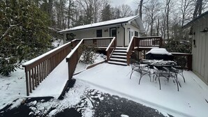Ein mit Schnee bedeckter Balkon mit Gartenmöbeln, eine Holztreppe, die zu einem Haus führt, und ein umzäunter Garten.