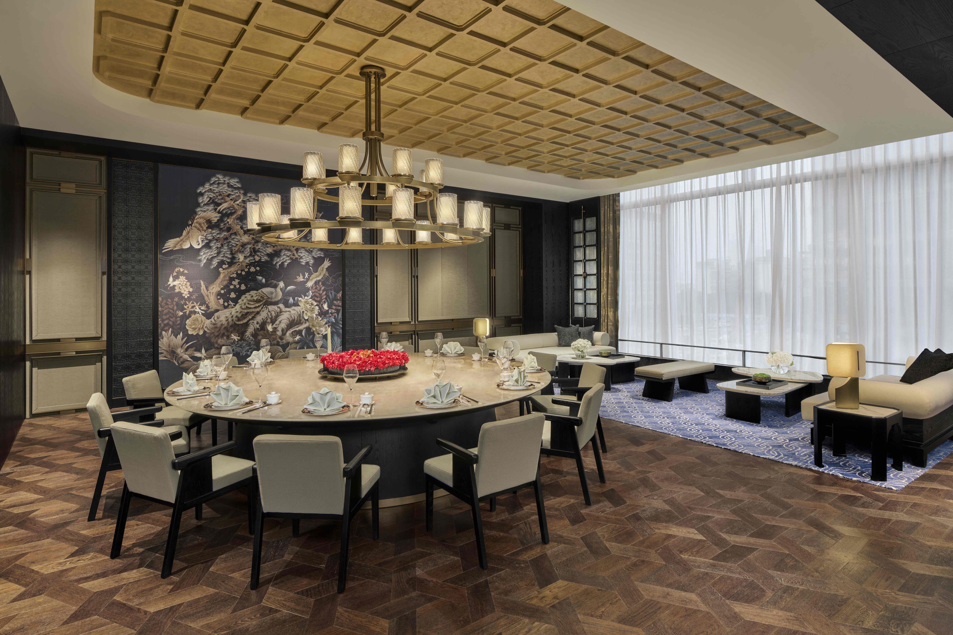 A modern dining area with a round table set for six, surrounded by chairs. A chandelier hangs above, and a large window with sheer curtains is visible in the background.
