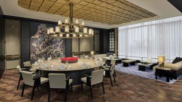 A modern dining area with a round table set for six, surrounded by chairs. A chandelier hangs above, and a large window with sheer curtains is visible in the background.