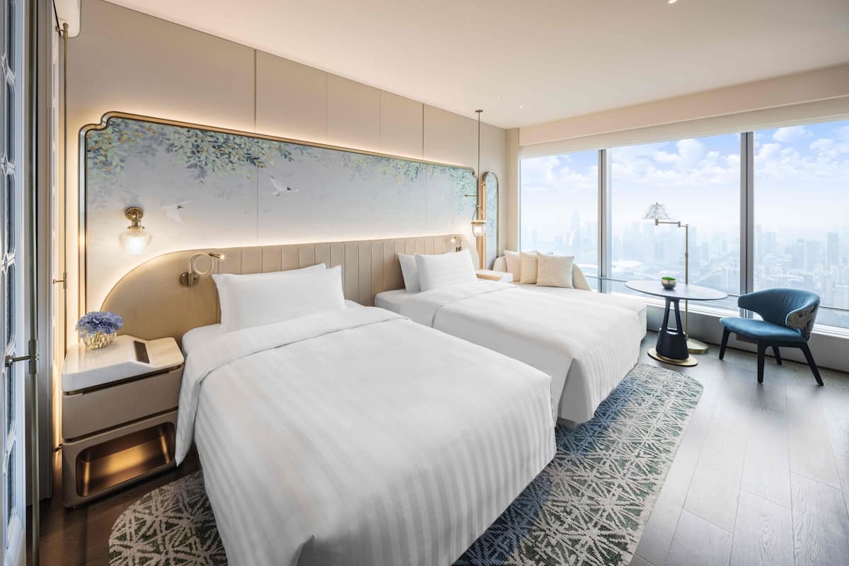 A modern hotel room with two beds, a large window with a city view, a desk with a lamp, and a chair.