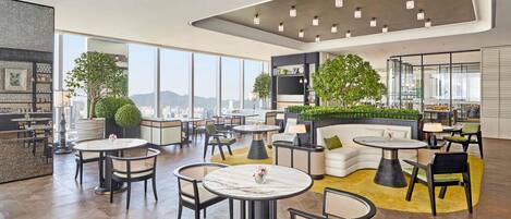 Amplia sala de estar con diversas opciones de asientos, incluyendo mesas redondas y sillas, un sofá y una zona de bar con vistas a la ciudad.
