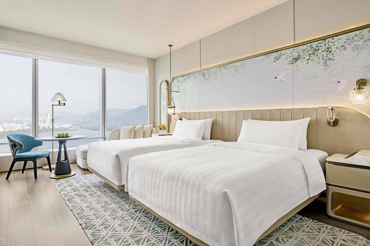 A hotel room with two beds, a view of the city, a round table with a lamp, and a large window.