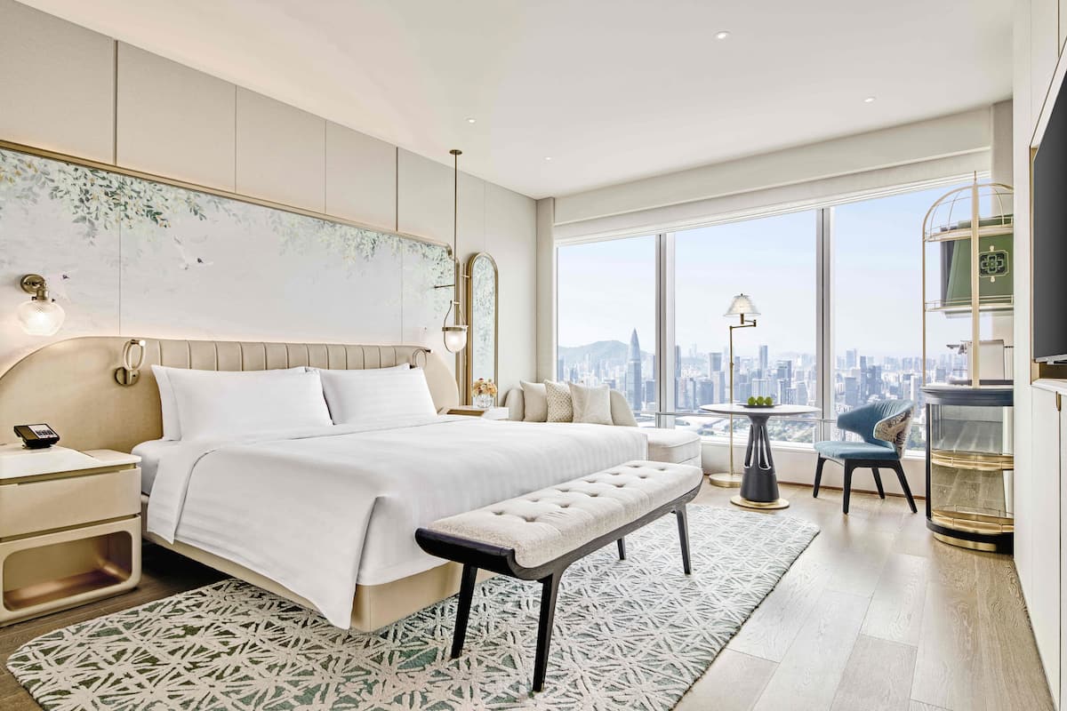 A modern hotel bedroom with a large bed, a bench, a nightstand, a side table, a chair, a round table, and a large window offering a city view.