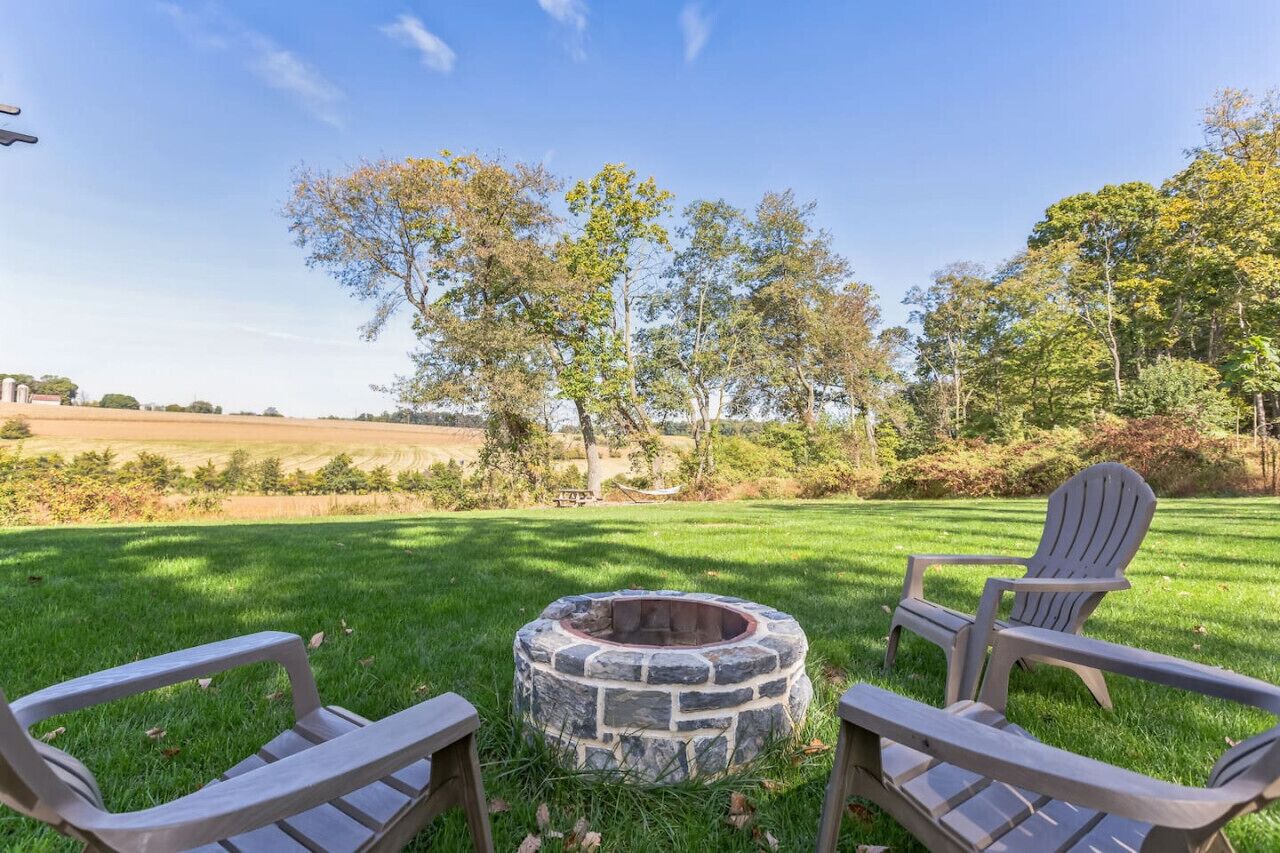 A stone fire pit surrounded by Adirondack chairs in a grassy outdoor area with trees and a field in the background.