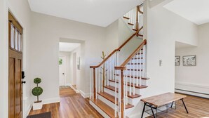 A spacious hallway with wooden flooring, a wooden staircase with white railings, and a small table with a wire basket.