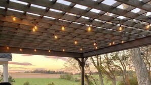 Un jacuzzi bajo una pérgola de madera con luces colgantes, rodeado de césped y árboles.