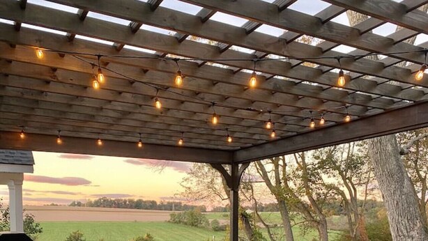 A hot tub under a wooden pergola with string lights, surrounded by a grassy yard and trees.