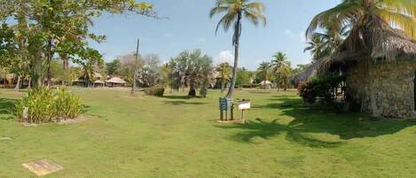 Un entorno tropical con chozas de techo de paja, palmeras y una zona cubierta de césped.