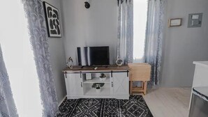 A room with a TV on a white cabinet, a small wooden side table, a patterned rug, and a window with sheer curtains. - De ja Blue (Saint George)