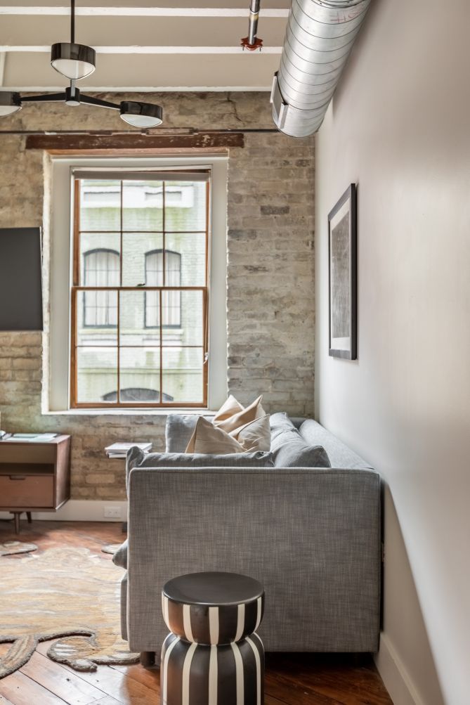 Un salon moderne avec un canapé gris, une petite table d’appoint ronde et une fenêtre donnant sur un immeuble.