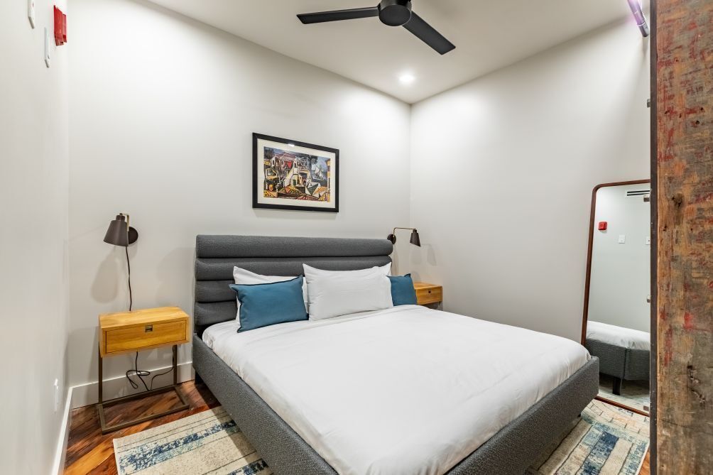 A bedroom with a bed, a ceiling fan, a nightstand, a lamp, a framed picture on the wall, and a mirror.