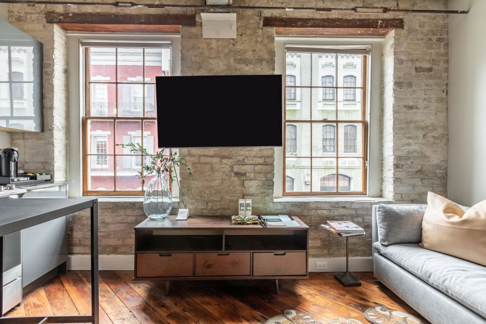 Een moderne woonkamer met zichtbare bakstenen muren, een groot raam, een flatscreen televisie aan de muur, een houten salontafel en een grijze bank.