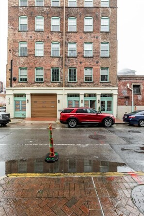 A multi-story brick building with a garage door, a red SUV parked in front, and a wet pavement reflecting the building.