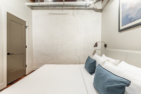 A bedroom with a white brick wall, a bed with white and blue pillows, a bedside table with a lamp, and a door on the left.