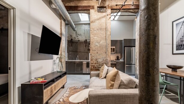 A modern living room with a large flat-screen TV, a sofa, and a coffee table. There is a kitchen area visible in the background. - Historic Charm Meets Luxury in This Central Warehouse Retreat (New Orleans)