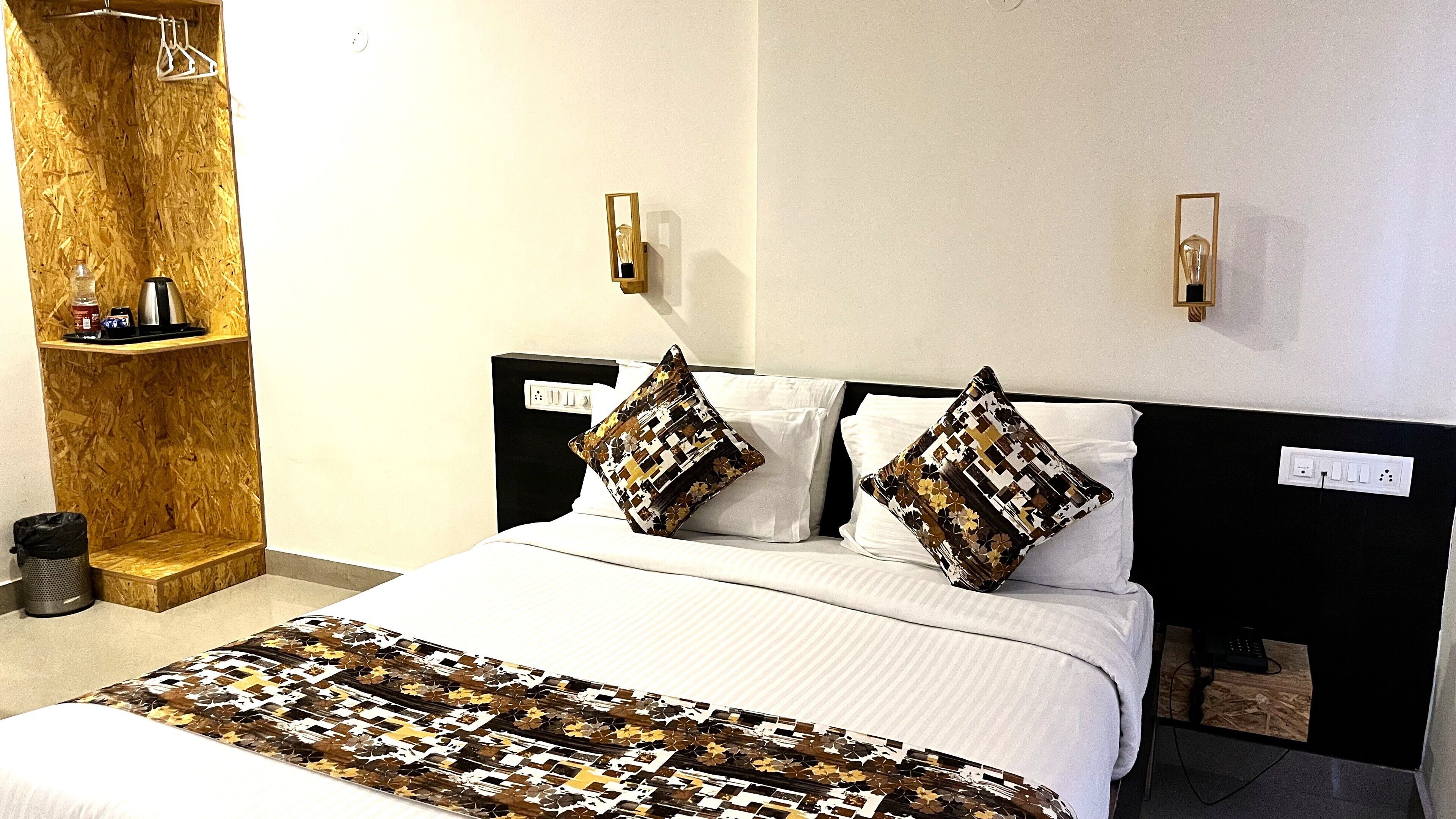 A hotel room with a bed, two bedside tables, a wooden shelf, and a trash bin.