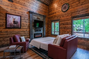 A log cabin interior with a fireplace, a bed, a red leather sofa, a red armchair, and a clock on the wall. - Romantic 1BR Hochatown Cabin w/ Hot Tub, Outdoor Fireplace & Soaking Tub (Broken Bow)