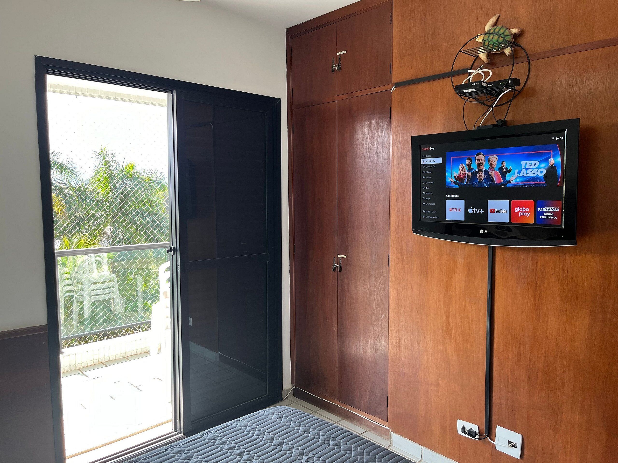 Una habitación de hotel con puerta de cristal corredera que da a un balcón, un televisor de pantalla plana en la pared y un armario de madera.